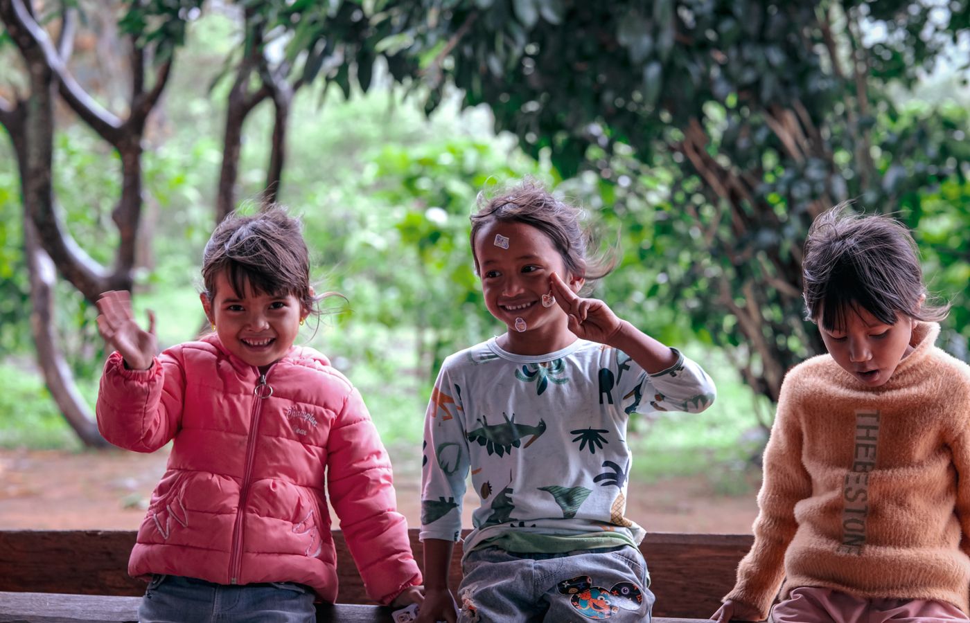 Three cheerful children sitting on a bench in a lush outdoor setting. They are smiling and interacting joyfully.