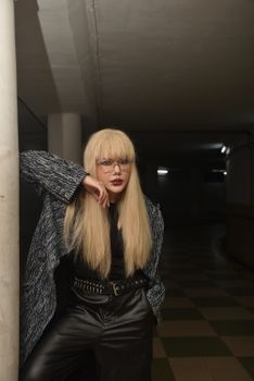 Stylish woman with long blonde hair poses confidently in a dimly lit underground location.