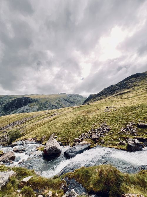 Free Scenic view of the Lake District with rocky stream and rolling hills under a cloudy sky. Stock Photo