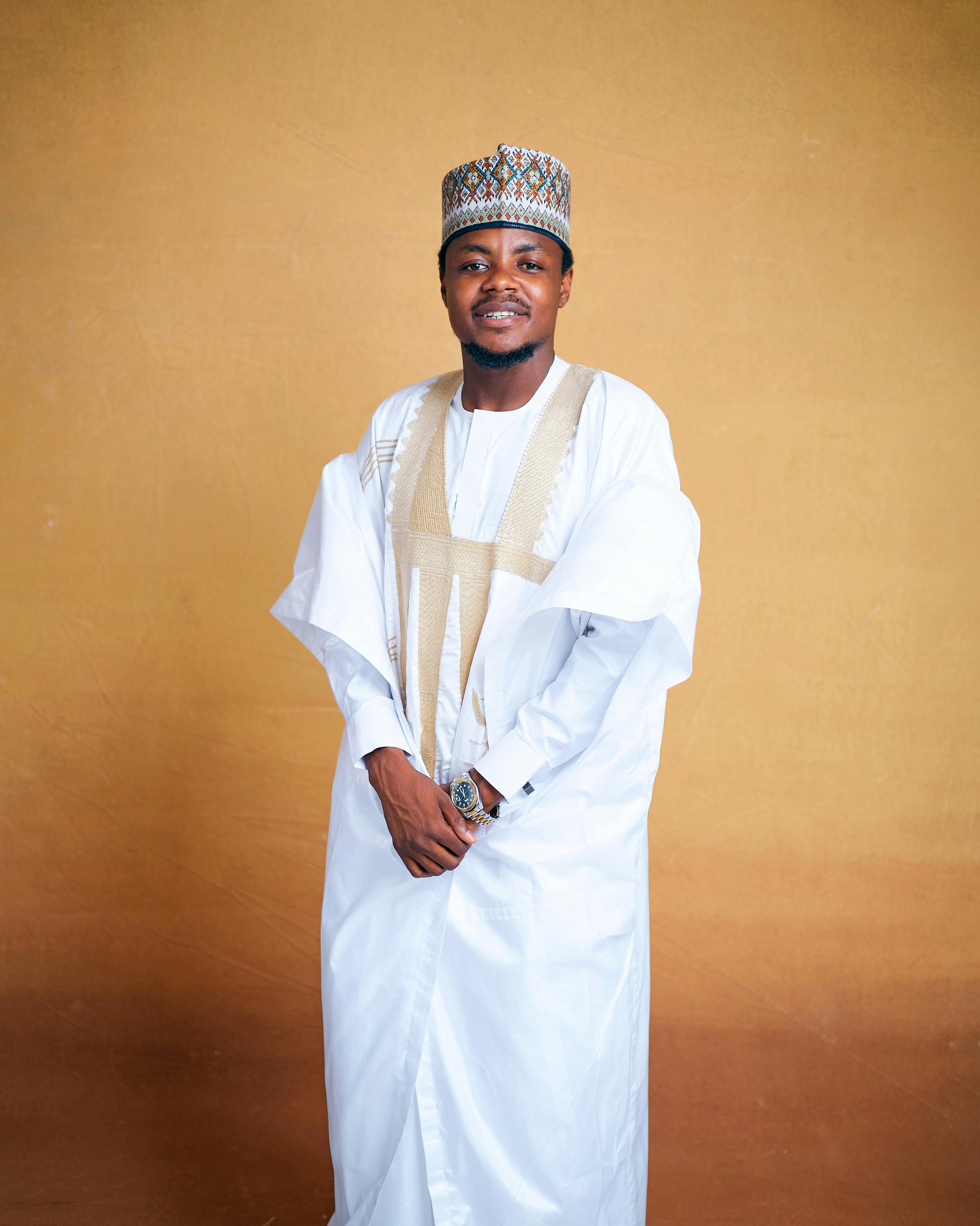 Man in classic Nigerian attire smiling indoors against brown background.