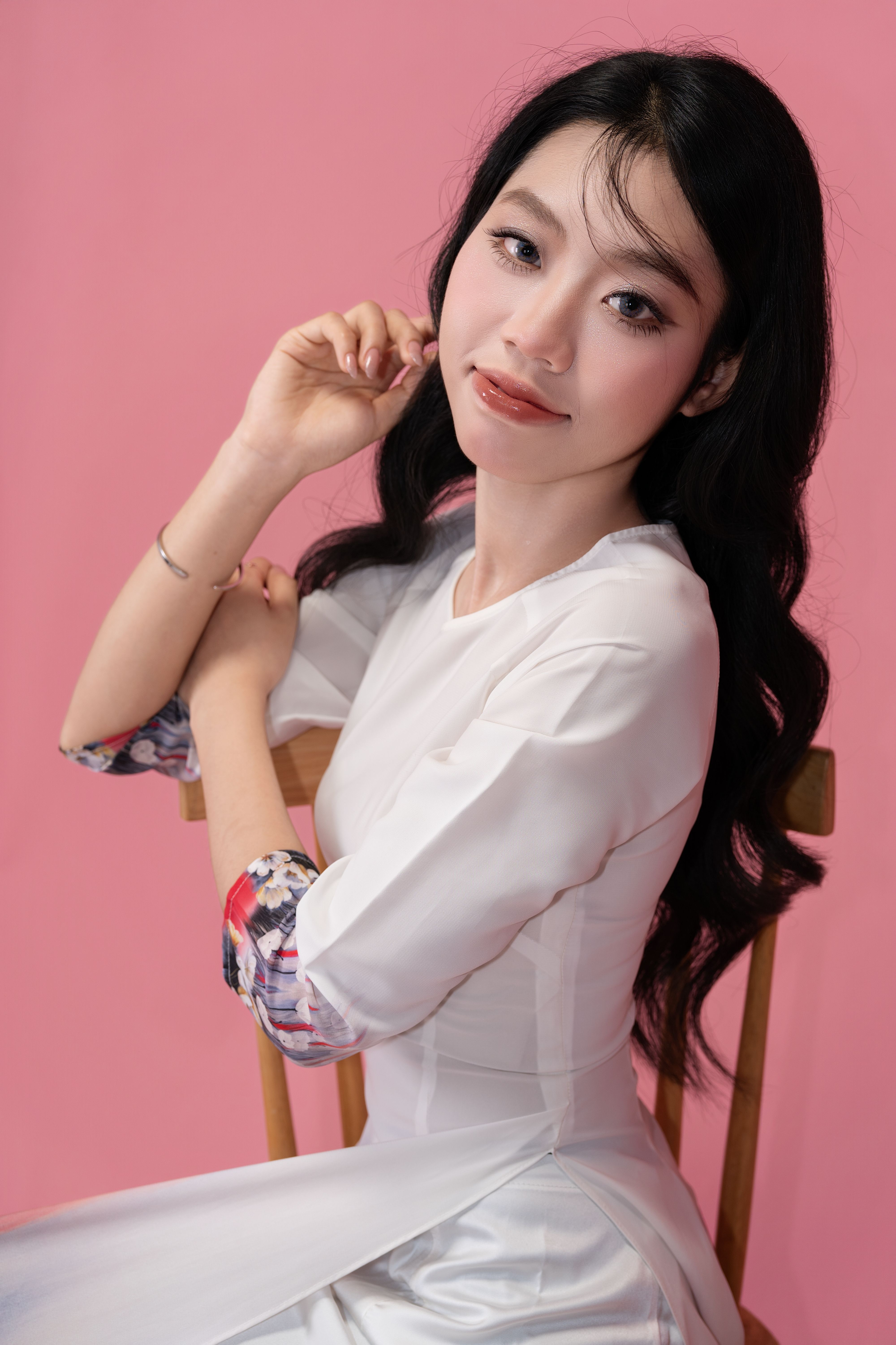 Woman in traditional white dress with floral accents poses against a pink backdrop.