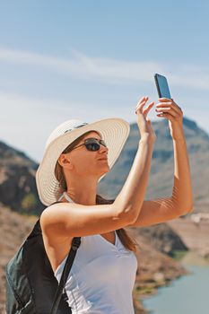 Young woman in sunglasses and hat capturing a moment with smartphone in a picturesque mountain setting.