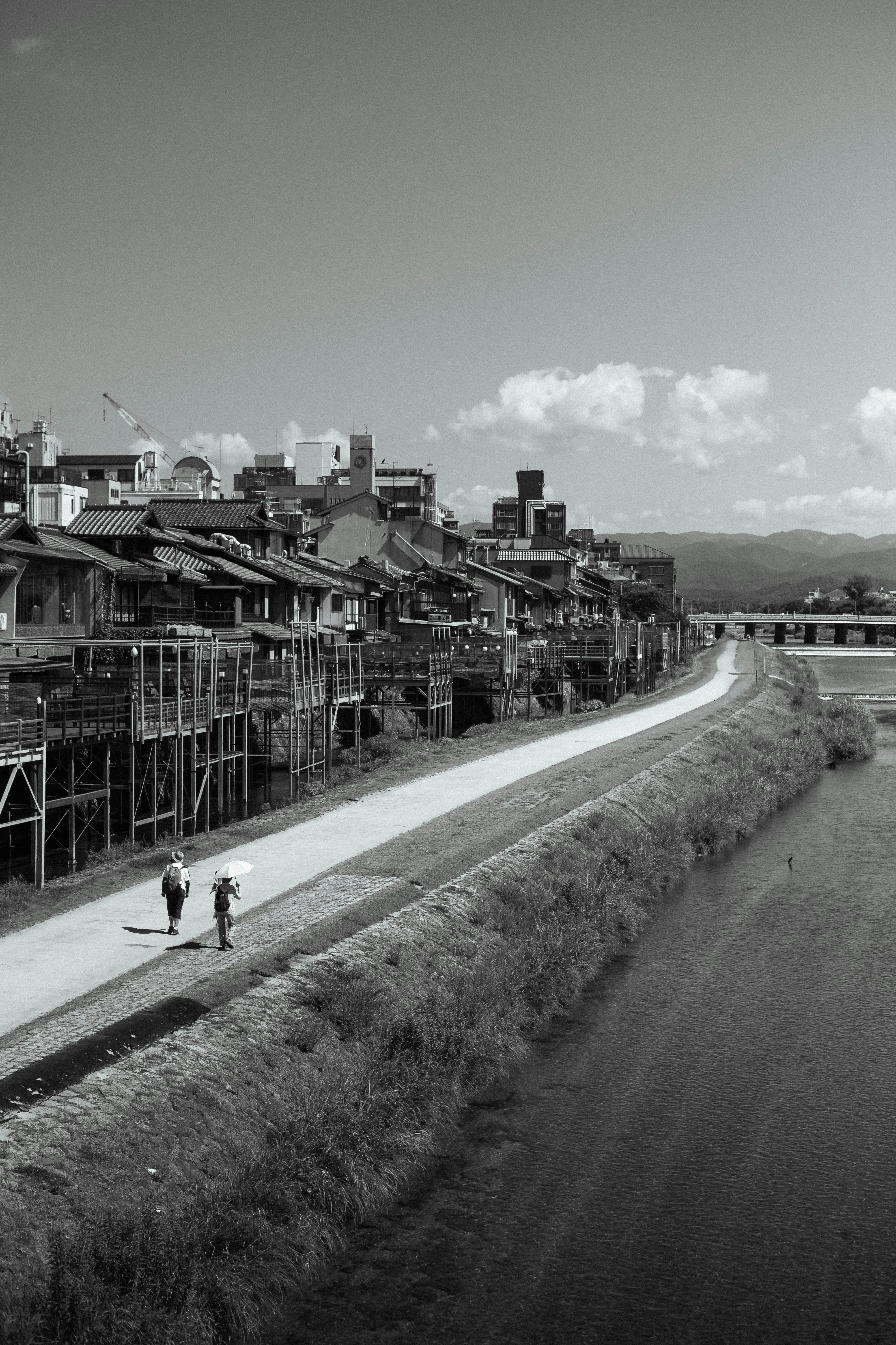 Atmospheric black and white photo of riverside promenade with two people walking in an urban setting.