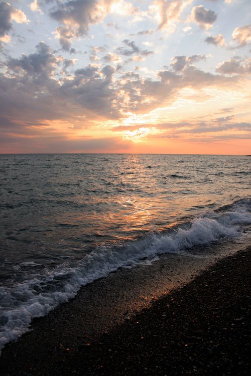 Free A beautiful sunset over a calm ocean with waves gently hitting the shore under a cloud-filled sky. Stock Photo