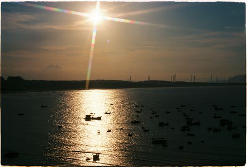 Free Golden sunset reflecting over calm sea with silhouetted boats and distant wind turbines. Stock Photo