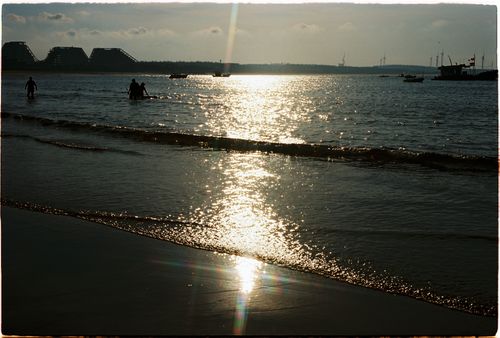 Free Silhouetted figures wade in water as the sun sets over a calm beach. Stock Photo