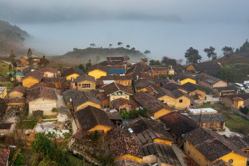 Free Charming aerial shot of a rustic village surrounded by mountains and mist, capturing a serene ambiance. Stock Photo