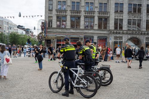 Free Three police officers on bicycles patrol a busy city square with people walking and historic buildings in the background. Stock Photo