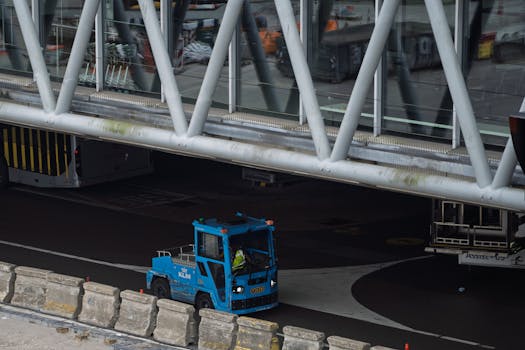 Blue airport ground vehicle moving under a jet bridge at a busy terminal.