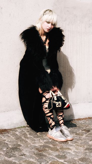 Fashionable woman in black fur coat and unique attire sitting outdoors. Edgy style.