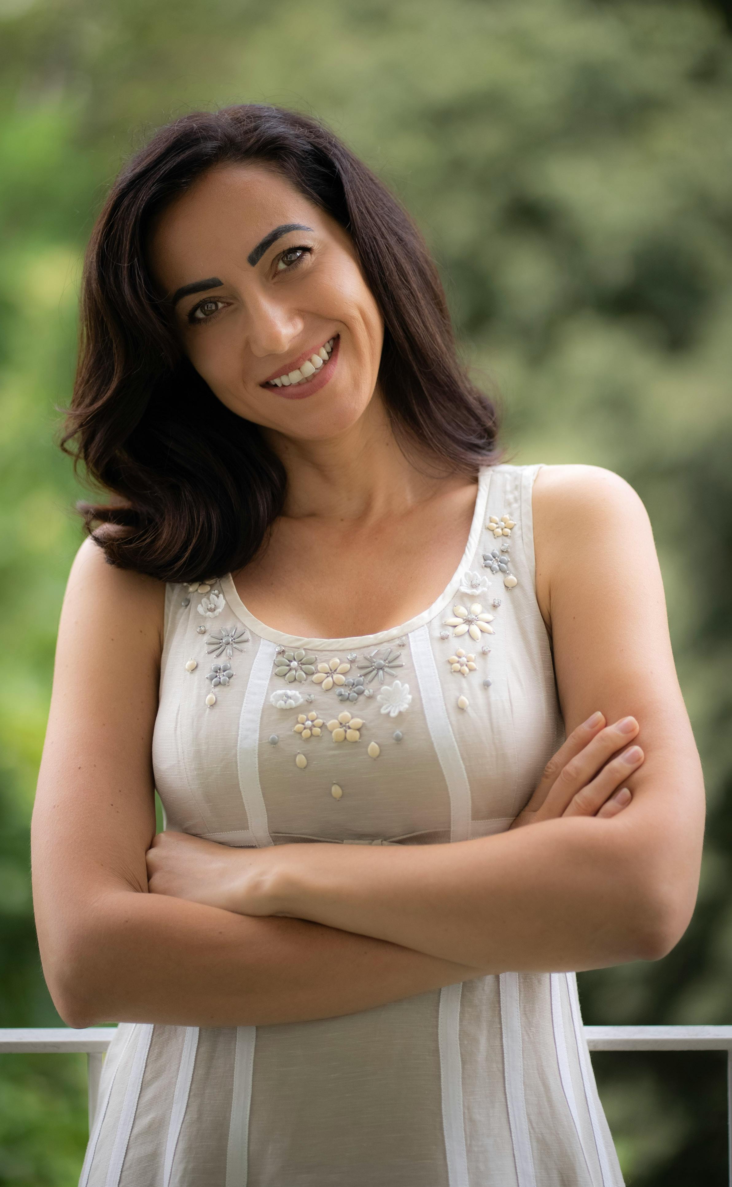 A smiling woman in a sleeveless dress posing outdoors, exuding elegance and confidence.