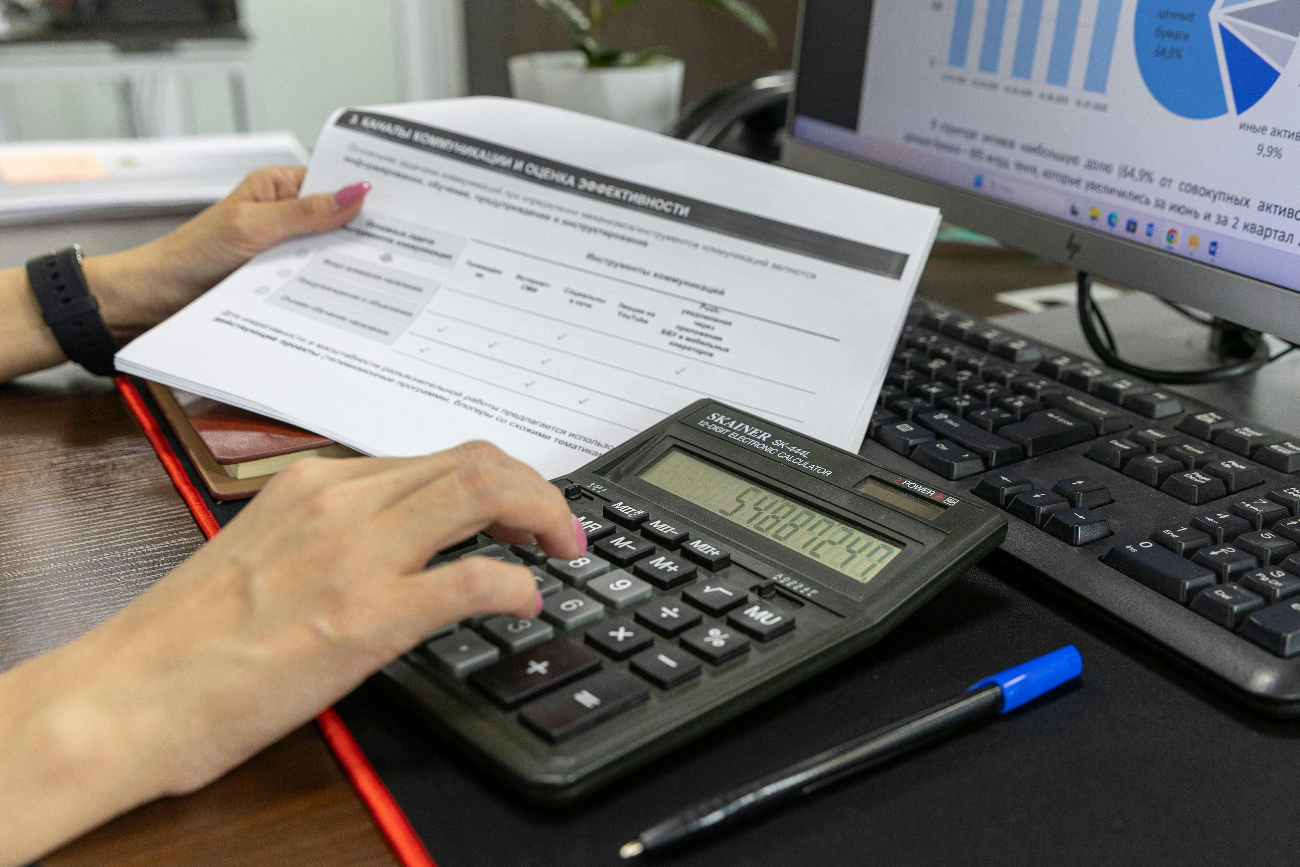 A person calculates financial data using a calculator and document, working at an office desk.