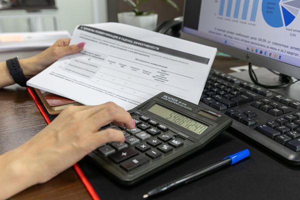 A hand on a desk calculator beside a printed form, a pen and a computer keyboard