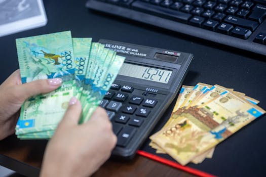 Hands counting Kazakhstani tenge while using calculator at desk.