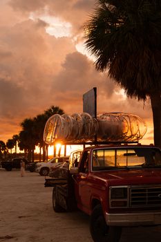 Red vintage truck parked near ocean at sunset with palm trees and dramatic sky.