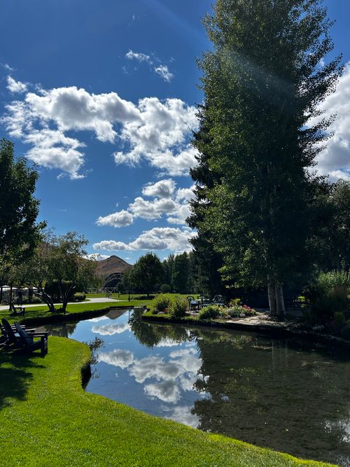 Free Serene park setting with a pond, lush trees, and scattered chairs under a bright blue sky. Stock Photo