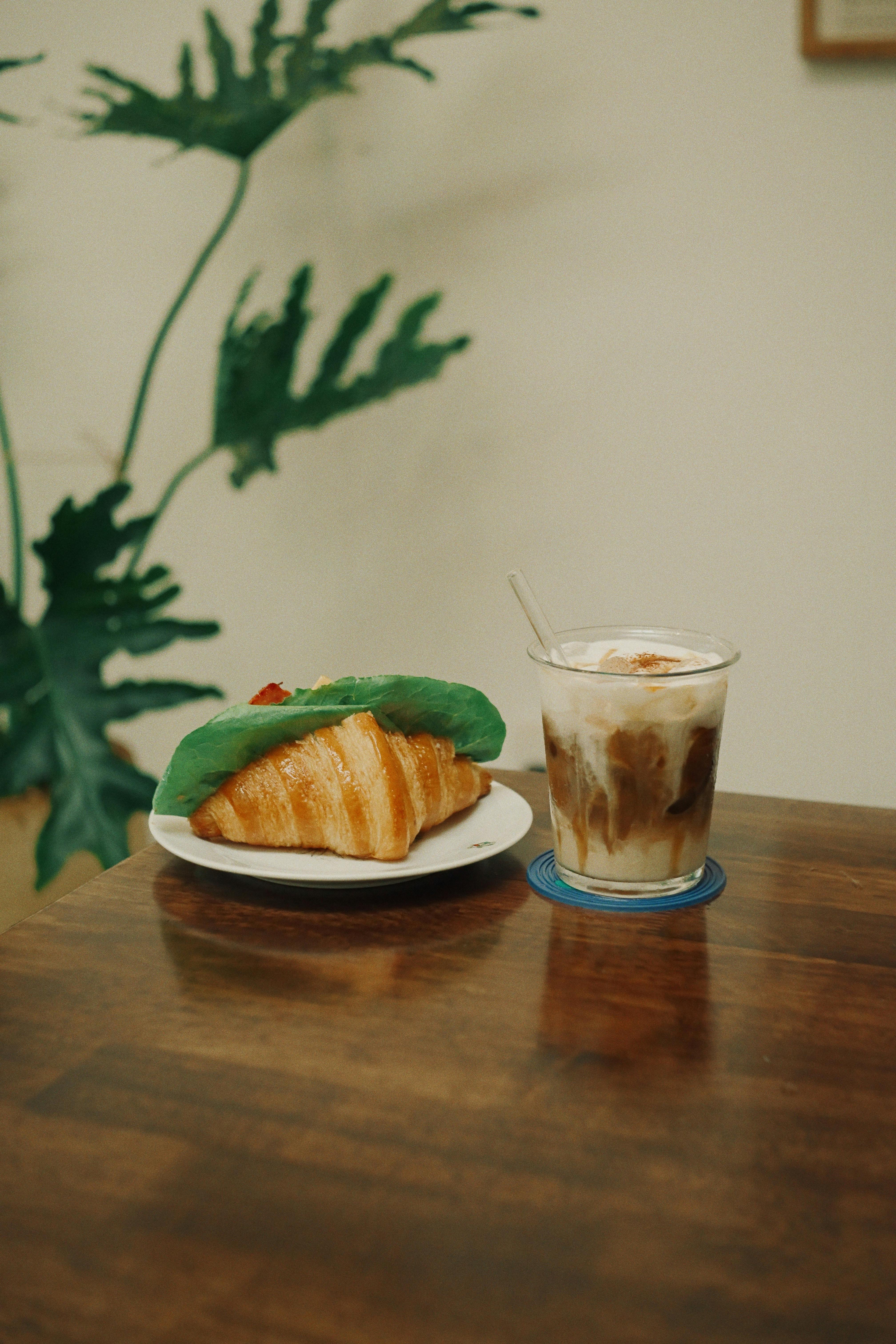 A fresh croissant and iced coffee placed on a wooden table with plant decor.