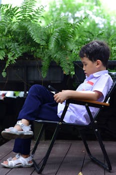 Young boy sitting outdoors on a chair, looking at a smartphone, surrounded by lush greenery.
