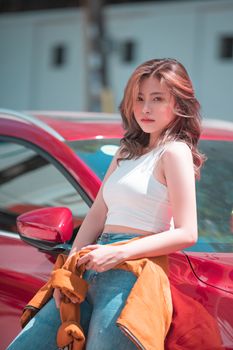 Stylish young woman in casual attire leans on red car under bright sunlight.
