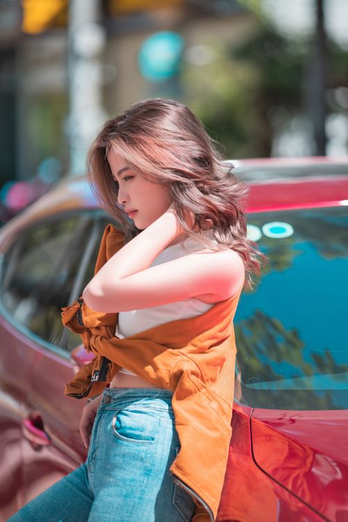Free Casual scene of a young woman in stylish attire leaning on a red car, bathed in warm sunlight. Stock Photo