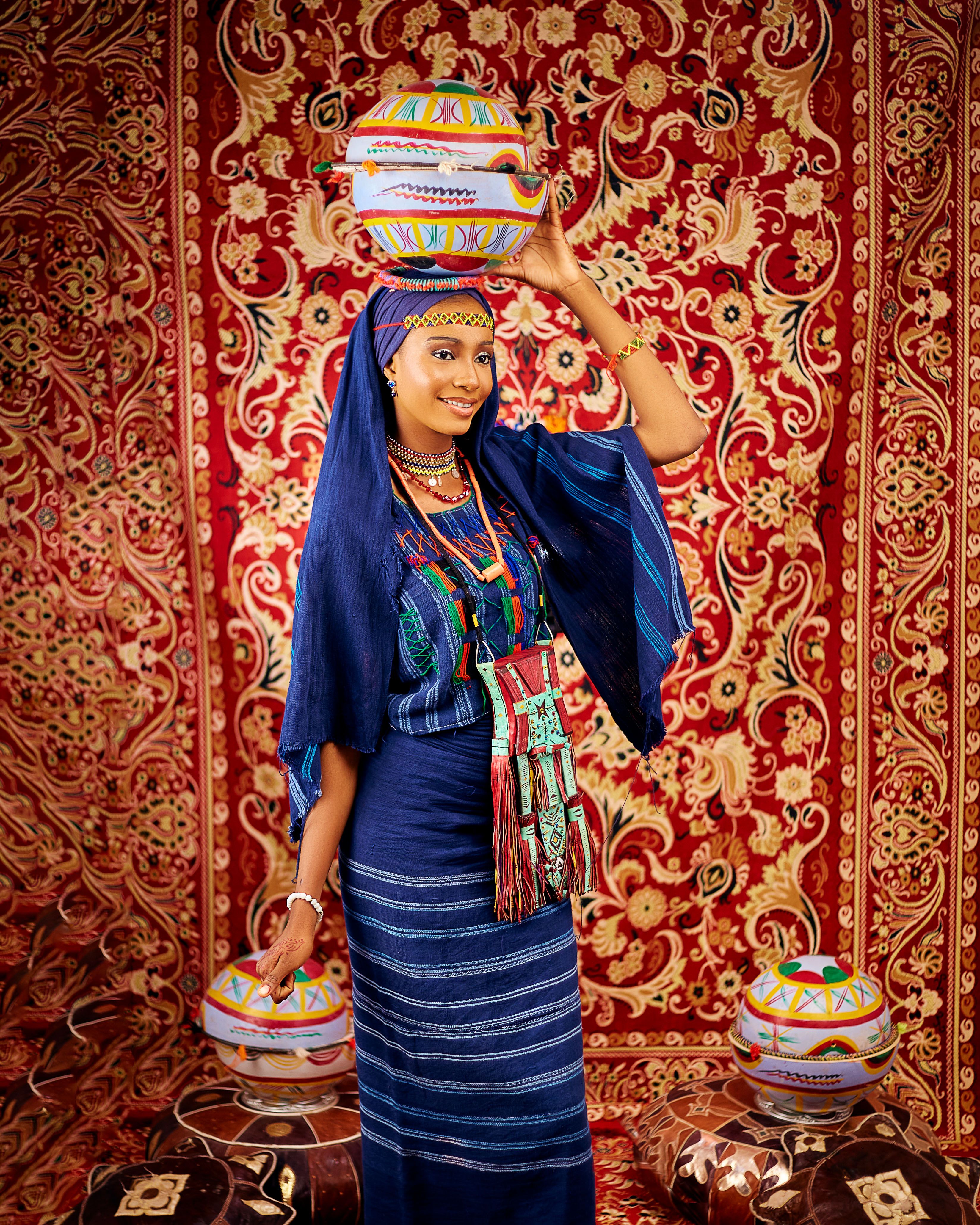 African woman in colorful traditional dress, balancing pottery on her head.