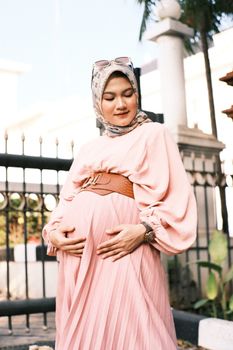 A pregnant woman in a pink dress and hijab, standing outdoors in a serene setting. - Fotografer Sidoarjo