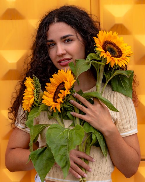 Free Young woman with curly hair poses with sunflowers against a vibrant yellow background Stock Photo