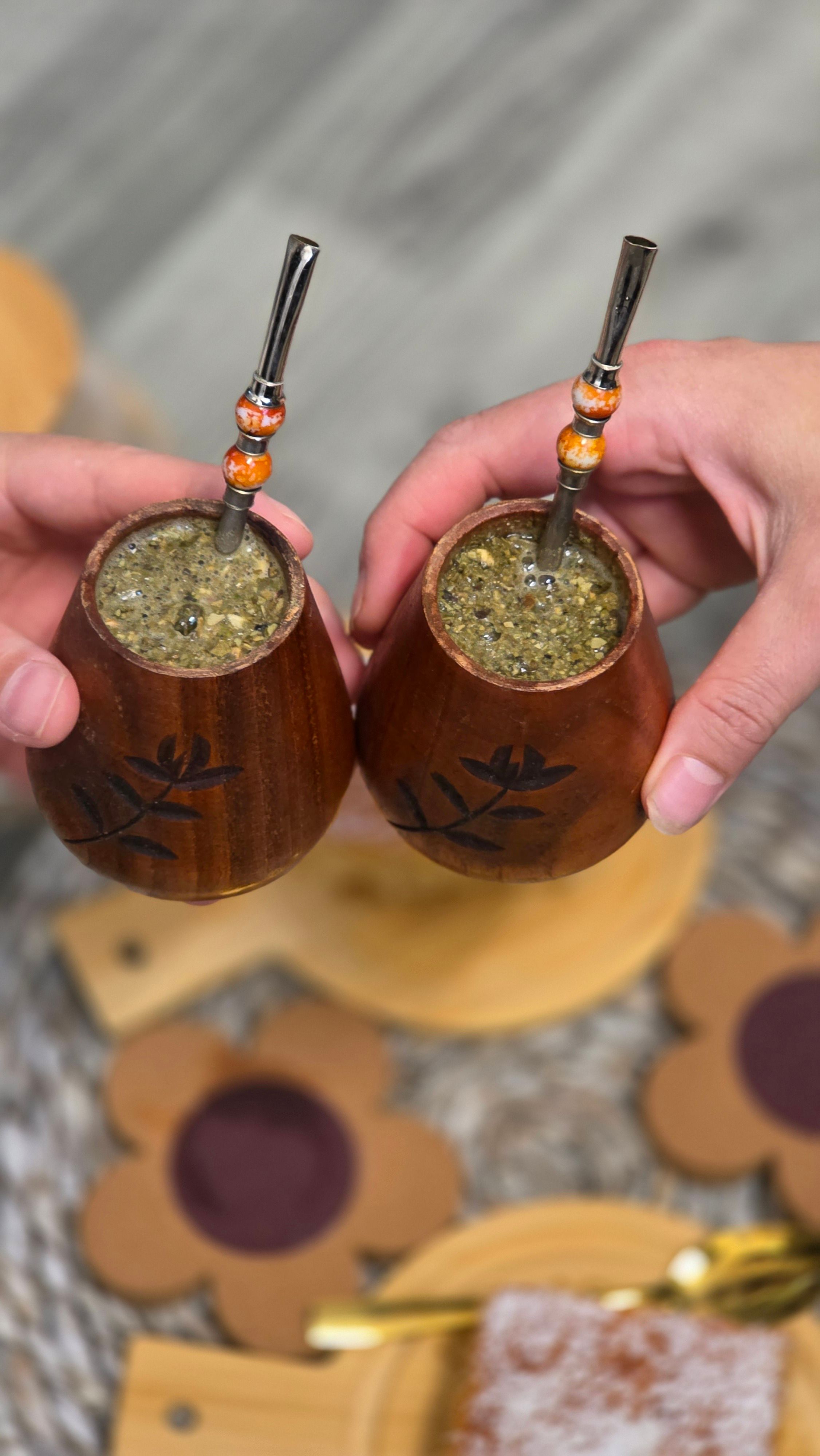 Two hands holding decorated wooden mugs of yerba mate with metal straws for a social beverage moment.
