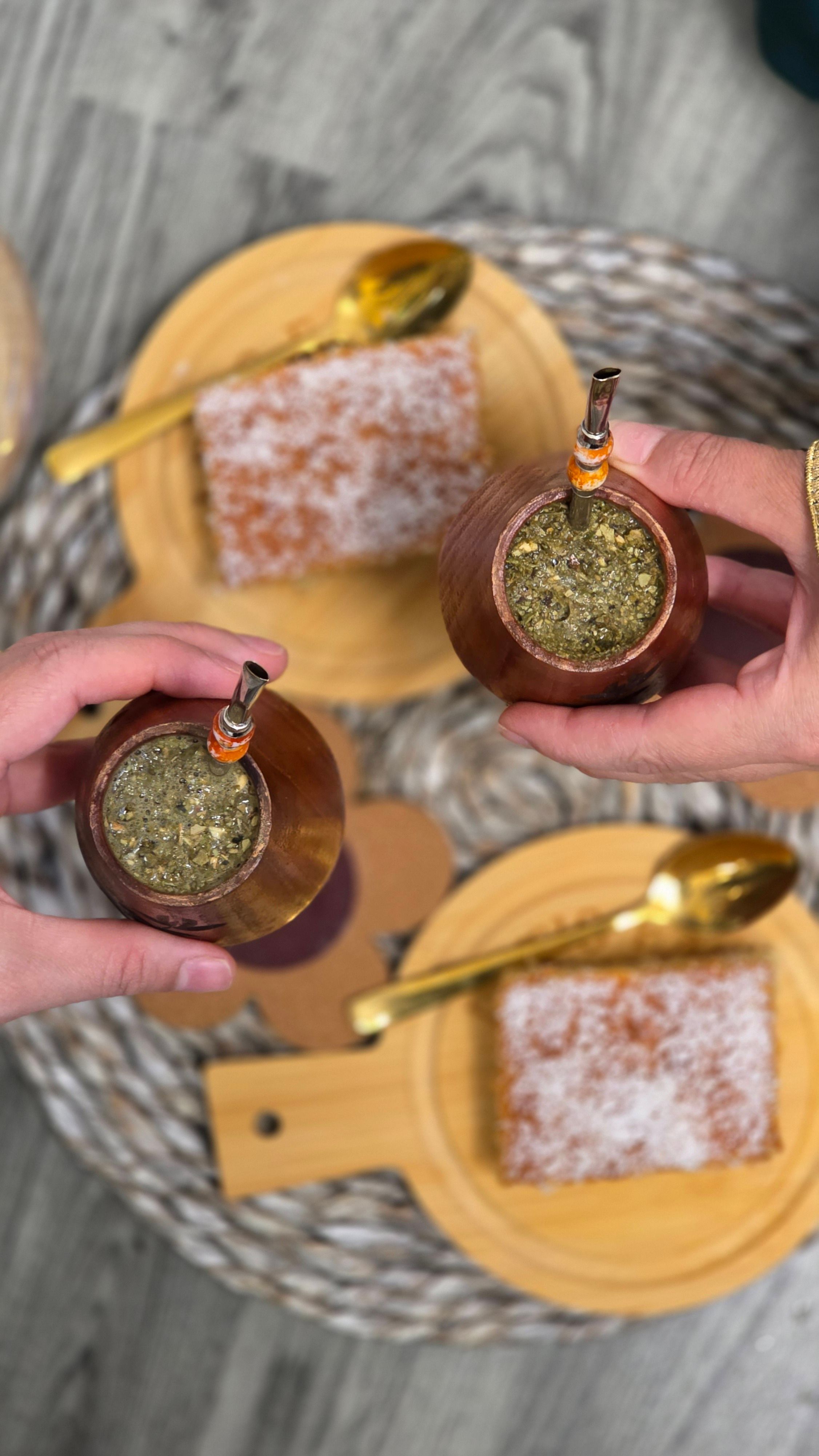 Two hands holding traditional mate cups over plates with cake, highlighting a casual social gathering.