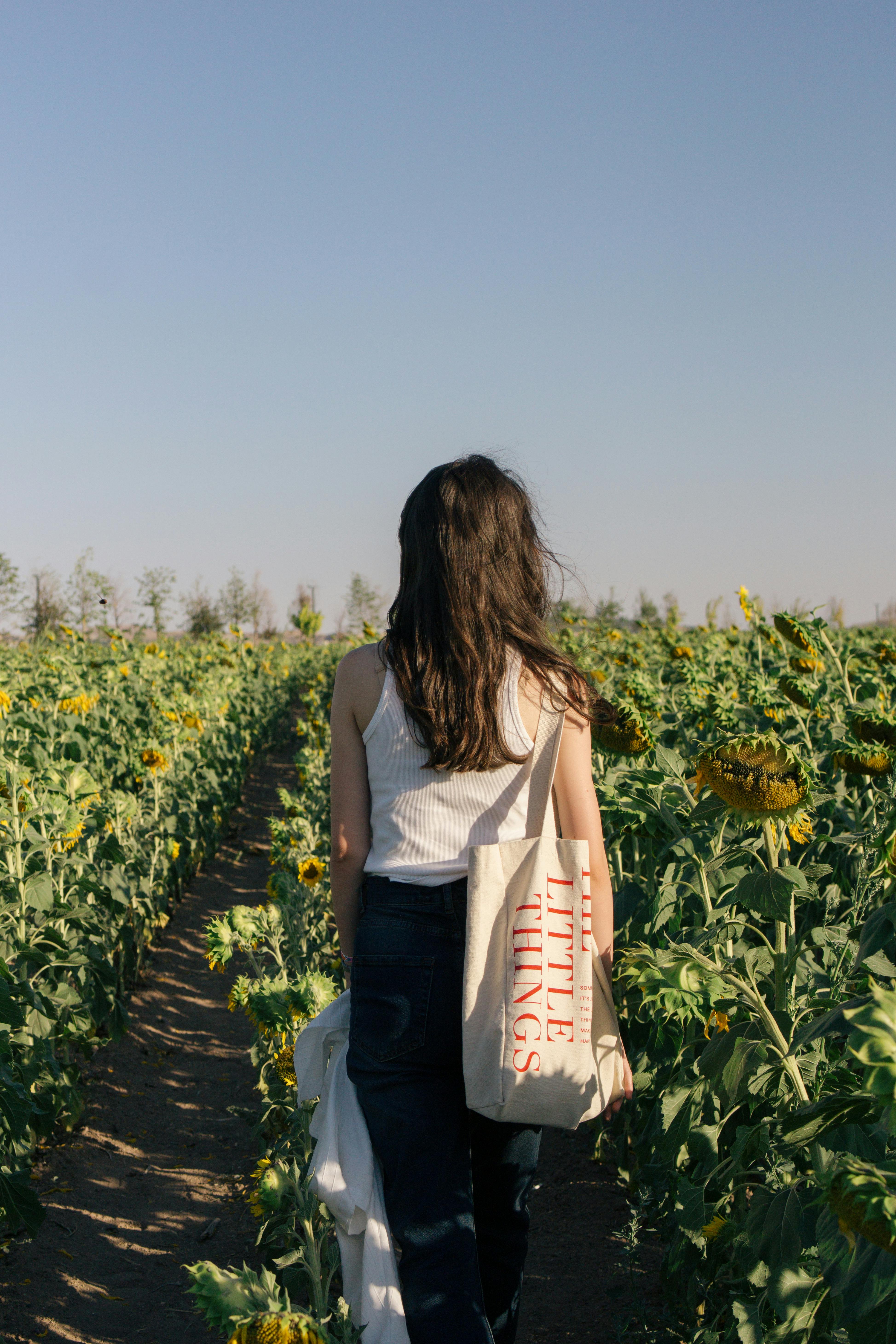 A woman walks through a sunflower field during daytime, carrying a tote bag.