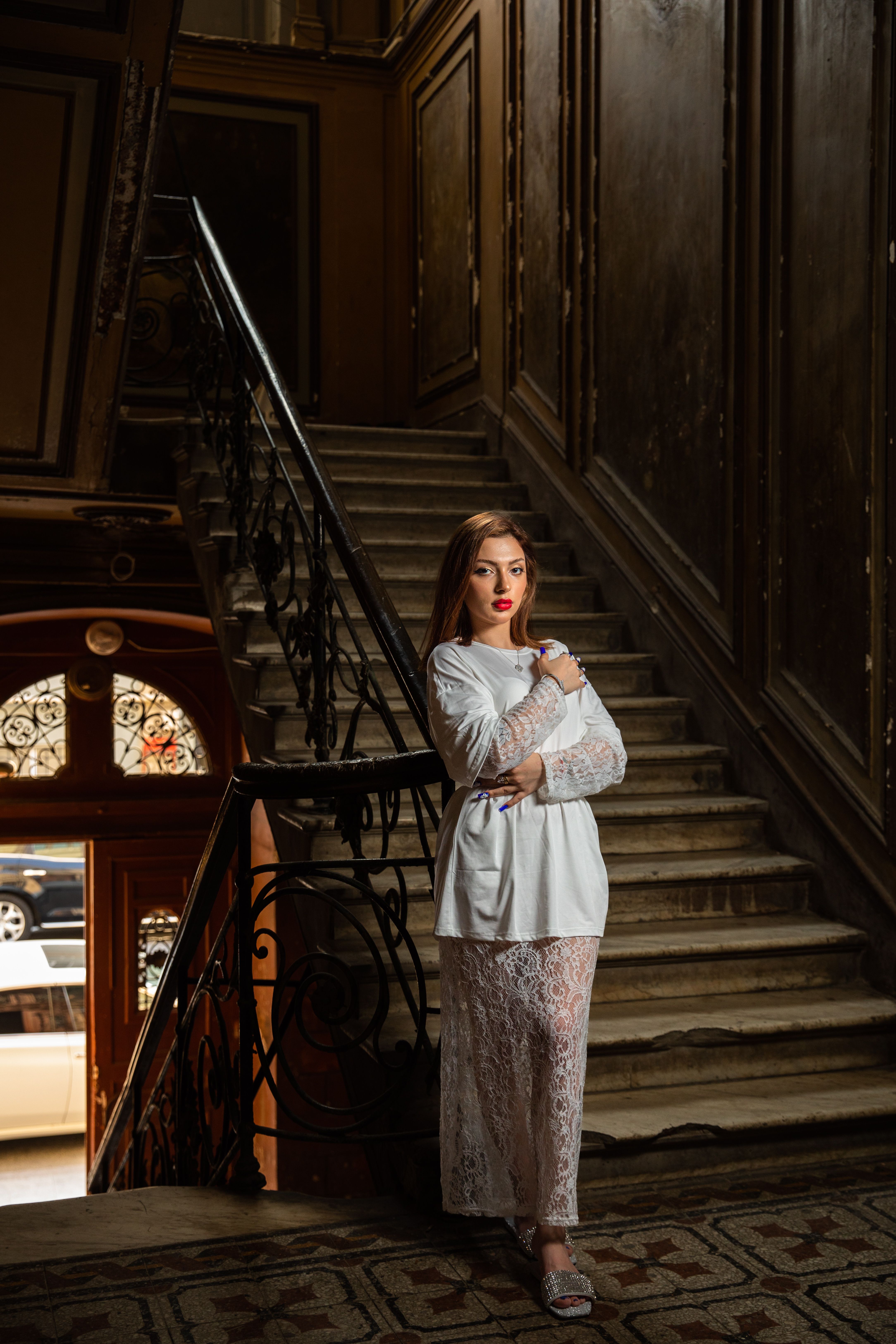 Stylish woman in white lace dress posing confidently on a grand staircase indoors.
