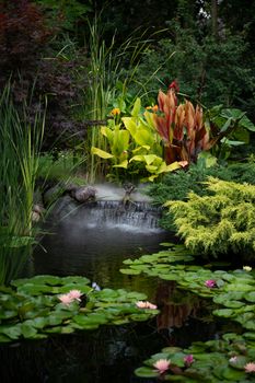 A tranquil garden scene featuring a small waterfall, vibrant greenery, and blooming lilies on a calm pond.