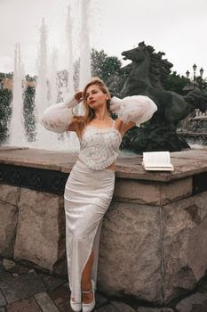 A woman in an elegant white dress poses creatively by a fountain with a sculpture in the background.