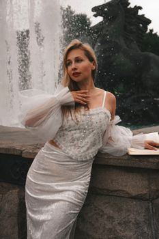 Stylish woman in white outfit posing elegantly by a fountain with horse sculpture.