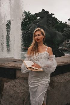 A woman in an elegant white dress reads a book beside a grand fountain statue.