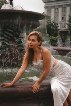 A woman in a white dress poses gracefully by a fountain in an urban setting.