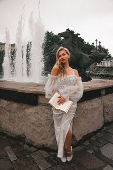 Woman in an elegant white dress standing by a fountain with an open book, exuding grace and tranquility.