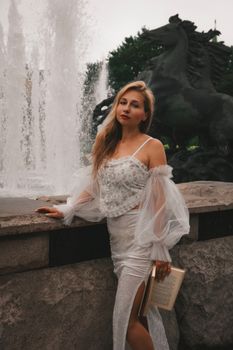 Elegant woman in white dress posing by fountain with horse statue.