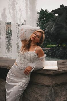 Elegant woman in white dress poses by a fountain, holding a book.