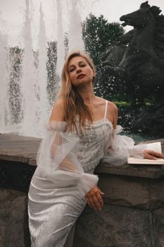 A woman in elegant attire poses confidently in front of a fountain and sculpture, creating a striking image.