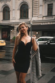 Stylish woman in a black dress enjoying a rainy day in the city, exuding confidence.