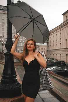 Stylish woman in black dress holding an umbrella on a rainy city street, exuding elegance.