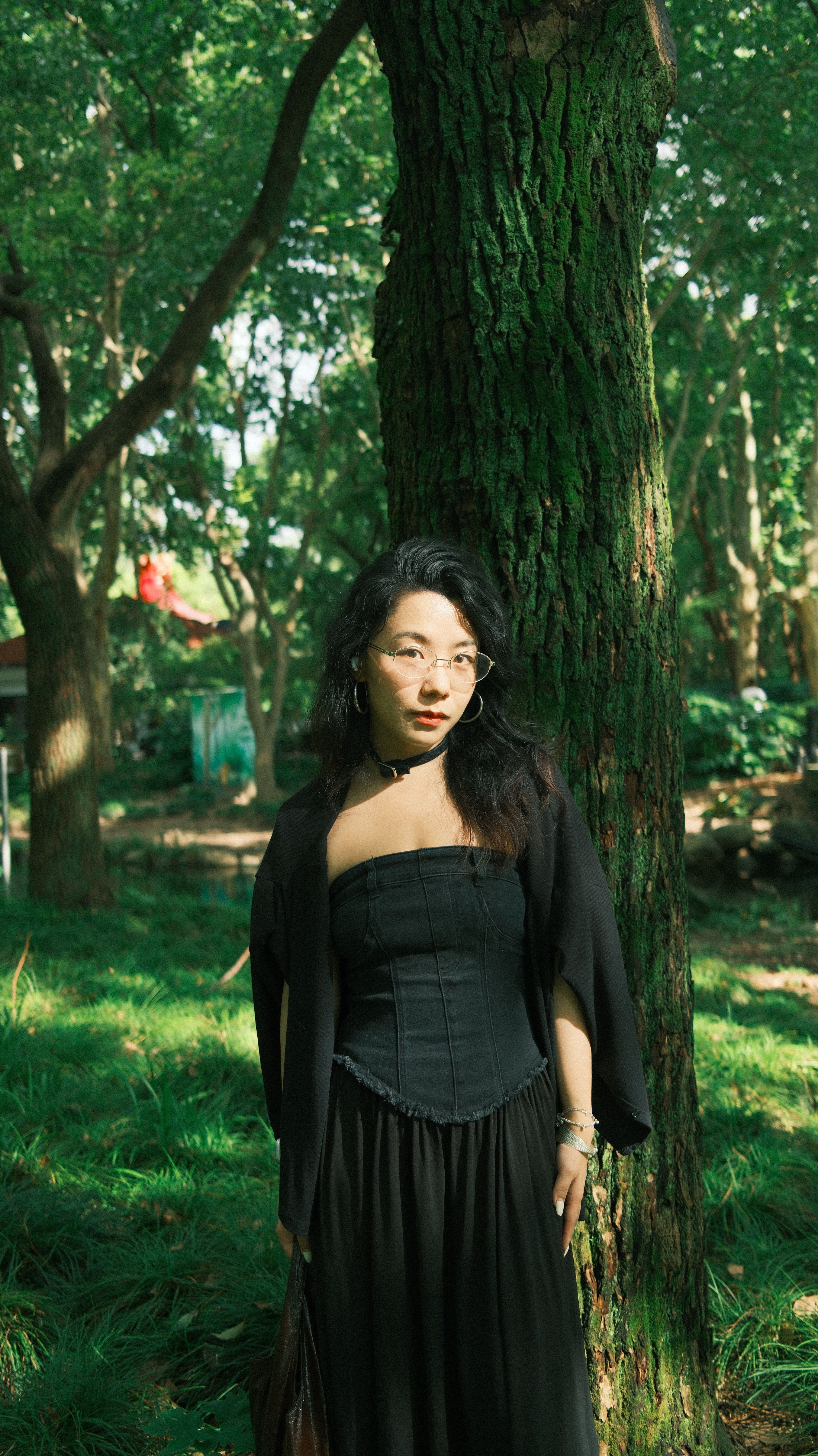 A woman in a fashionable black dress stands amidst lush green forest surroundings.