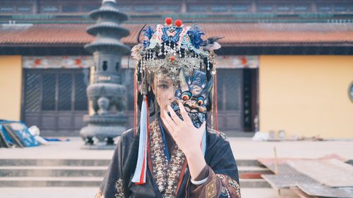 Free A person in traditional attire and ornate headpiece holds a mask in an outdoor cultural setting. Stock Photo