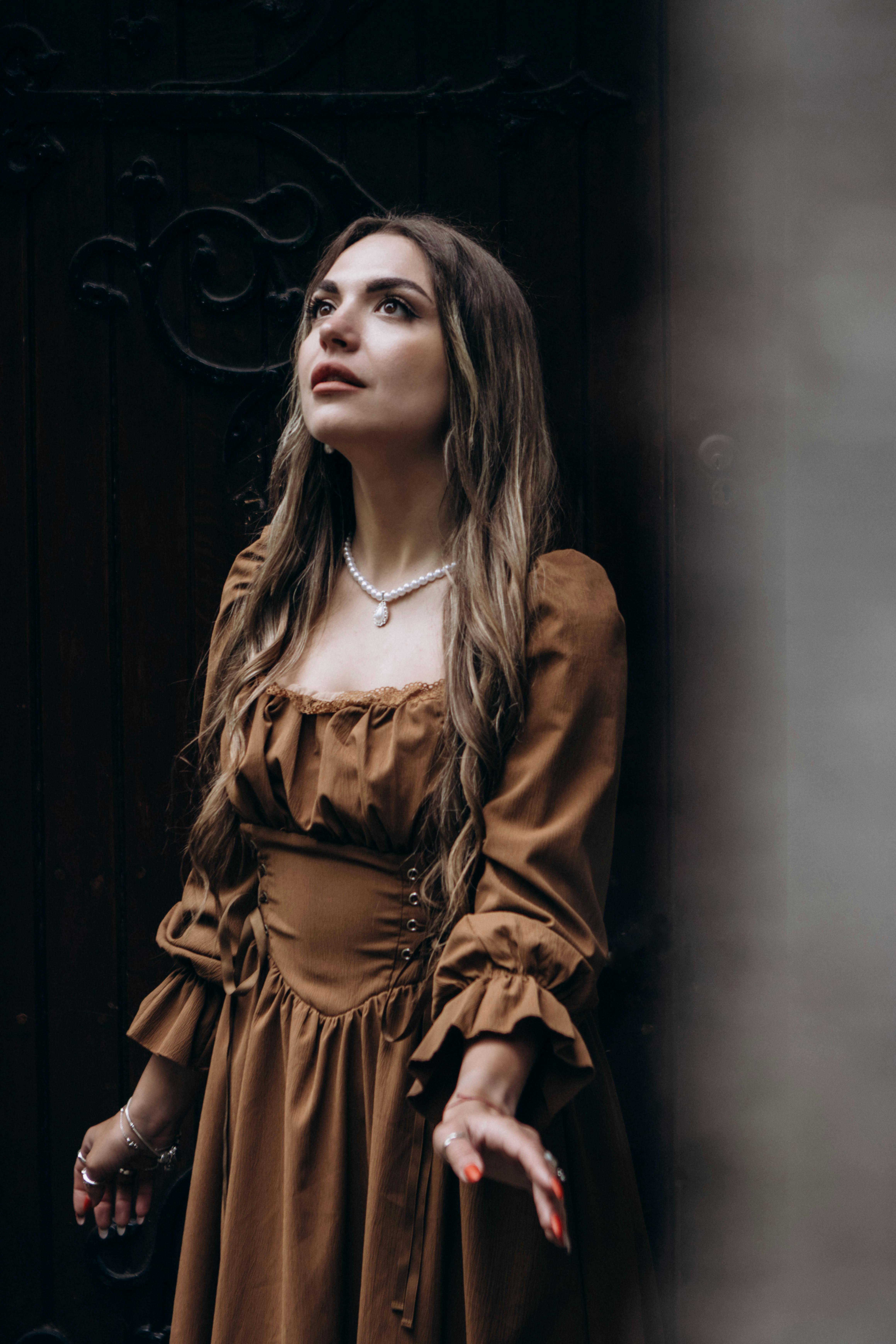 Free A woman in a vintage brown dress looks upward with a thoughtful expression. Stock Photo