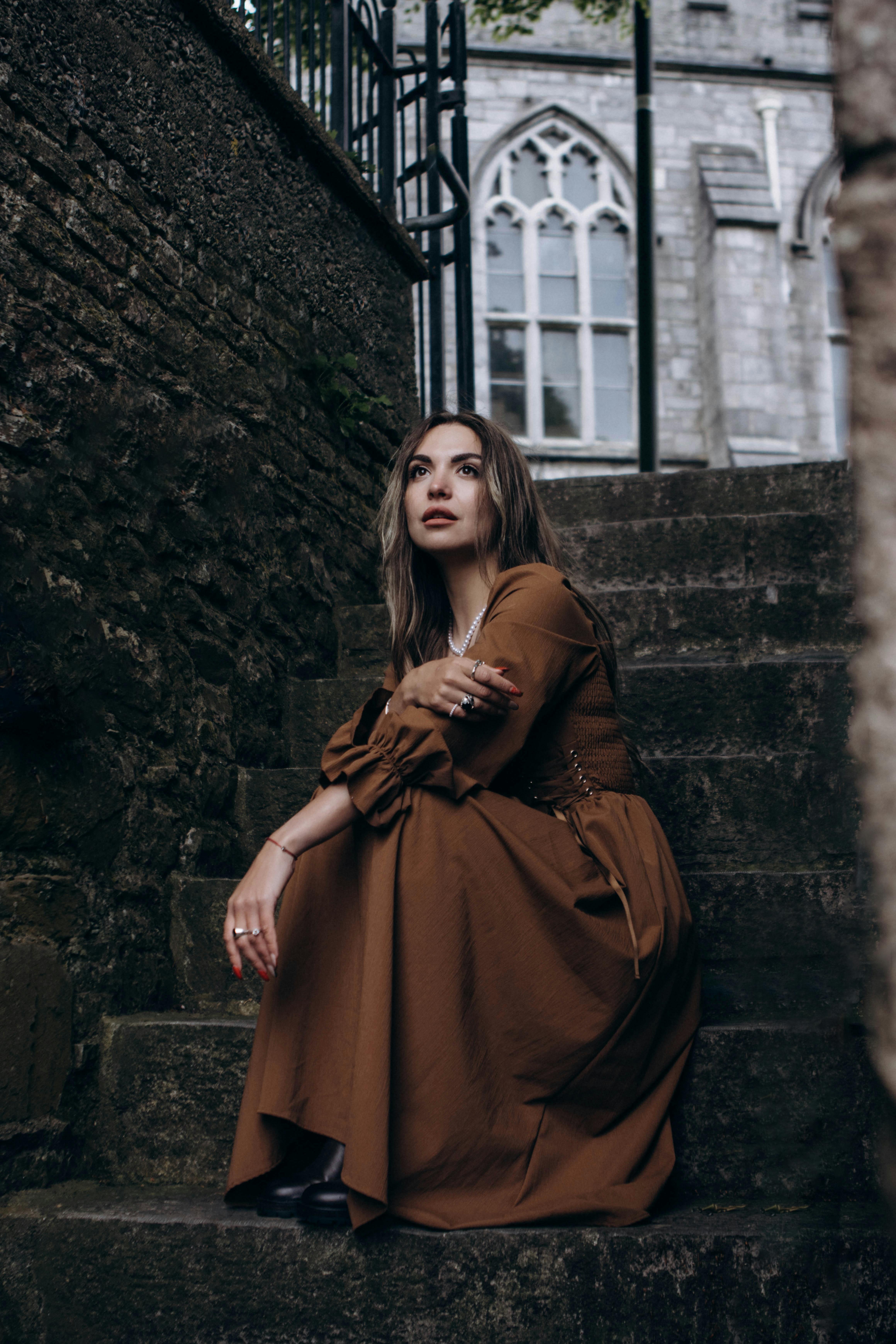 Woman in brown medieval dress poses on stone steps by historic building.