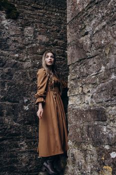 Woman in brown vintage dress standing by a stone wall outdoors. Rustic, elegant fashion.