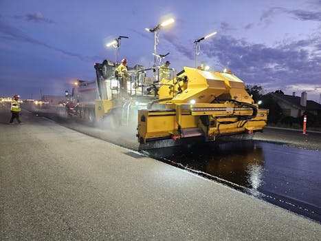 Workers operate heavy machinery to pave a road at twilight, enhancing infrastructure.