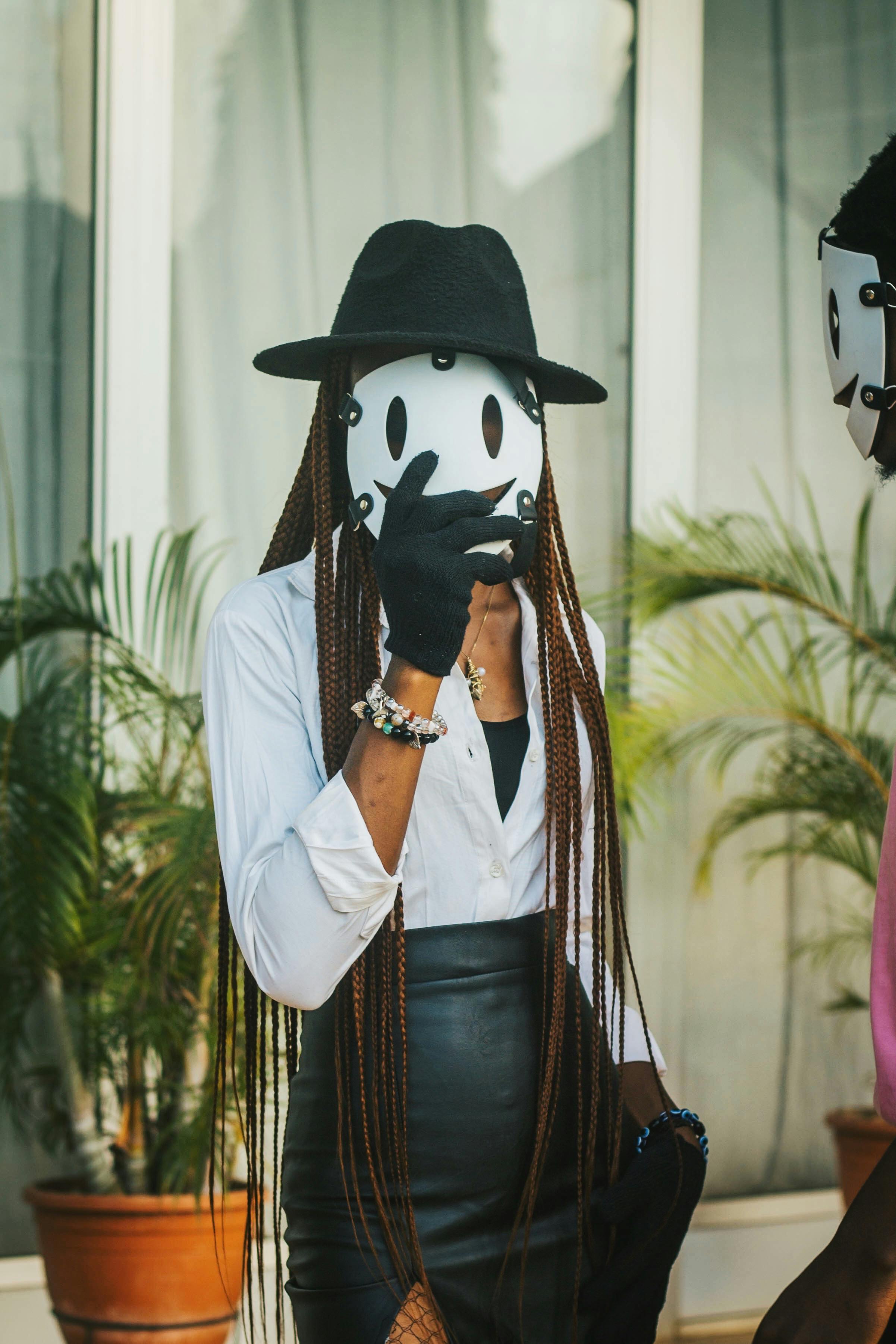 Woman in a mask with stylish outfit, black hat and gloves poses outdoors, exuding mystery.