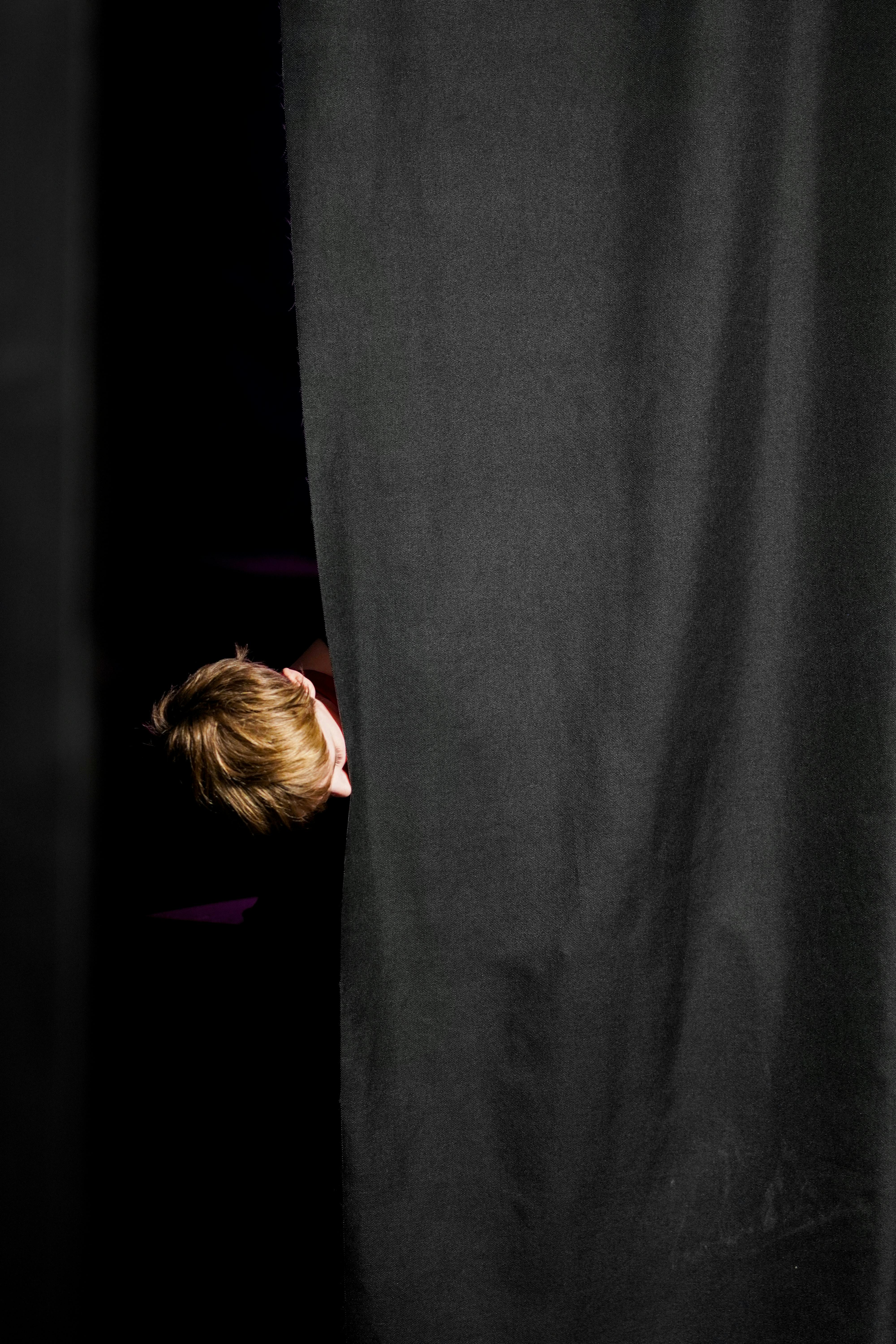 Kostenlos Eine junge Person lugt hinter einem großen schwarzen Vorhang hervor und erzeugt so ein Gefühl des Geheimnisvollen. Stock-Foto
